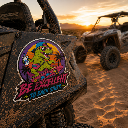 A colorful die-cut sticker featuring a cartoon green dinosaur wearing a blue cap and sunglasses, holding a drink and riding a pink skateboard, with the text 'BE EXCELLENT TO EACH OTHER' below it. The sticker is shown applied to the side of a black off-road vehicle in a desert setting at sunset, with another UTV in the background.